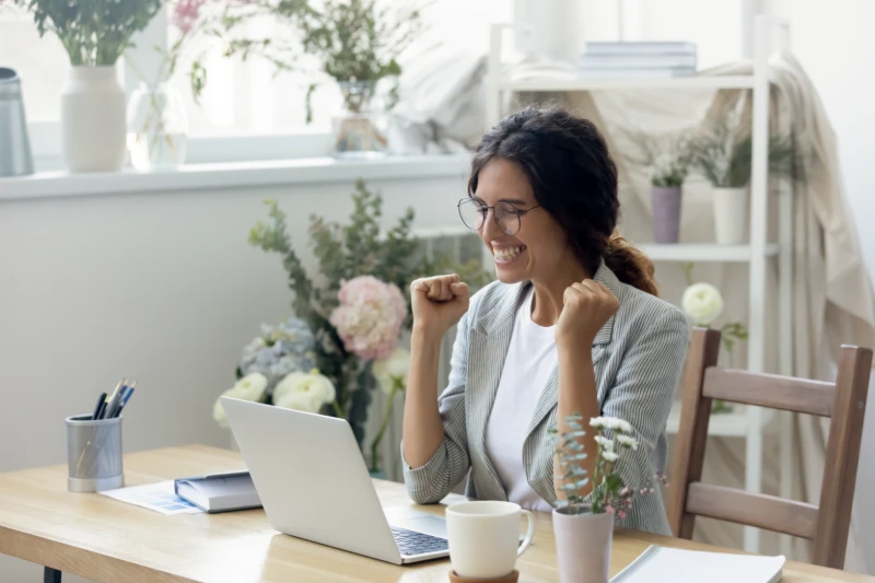 Home 45 A woman sits at a desk with a laptop, smiling and raising her fists in excitement. The room has plants and flowers, and sunlight streams through a window in the background.