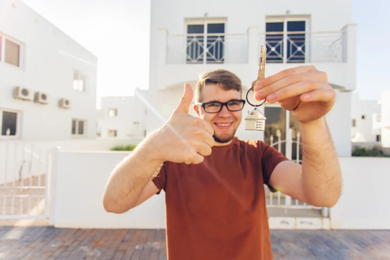 Home 43 A smiling man wearing glasses holds up a house key and gives a thumbs up, standing in front of a white residential building on a sunny day.