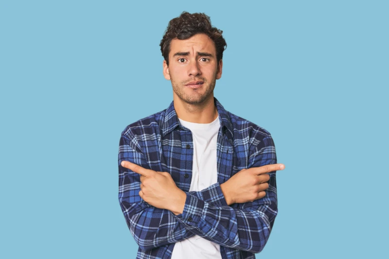 Home 42 A man in a blue plaid shirt stands against a light blue background, crossing his arms and pointing in opposite directions with both index fingers, looking confused or uncertain.