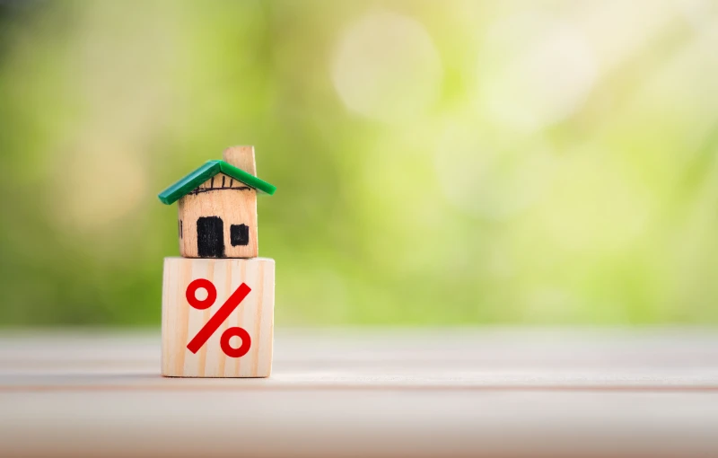 Home 44 A small wooden house figurine sits on top of a wooden block with a red percent symbol, against a blurred green and beige background.