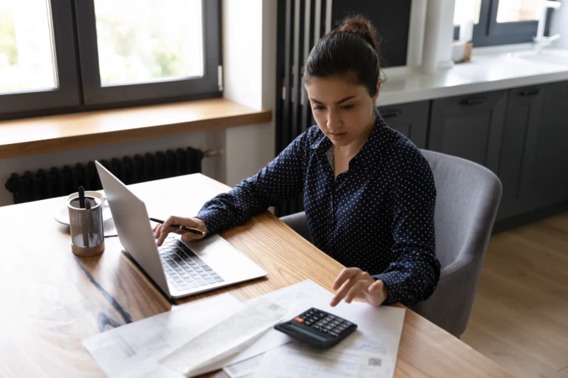 Home 40 A woman sits at a table using a laptop and calculator, surrounded by papers and a pen holder, appearing focused while working in a modern home setting with large windows.