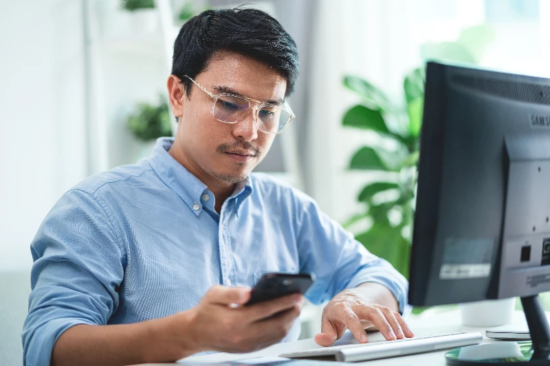 Home 18 A-man-wearing-glasses-is-sitting-at-a-desk-with-a-computer-monitor-and-a-cell-phone-He-is-looking-at-the-cell-phone-while-typing-on-the-computer